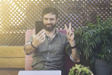 Çekici bir adam kendini üstünde hareket eden telefon fotoğraf çekimi ve barış hareketi gösteren şık giysiler içinde içerik. Sakallı hippi selfie terasta smartphone üzerinden yapmak