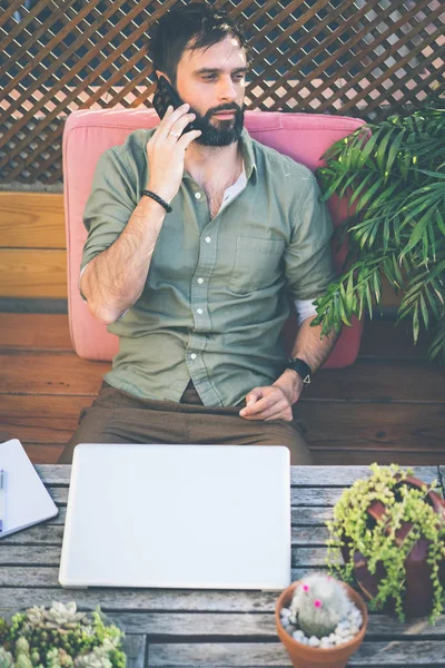Uomo hipster che lavora sul computer portatile e utilizzando il telefono cellulare mentre seduto sulla terrazza del caffè. Attraente uomo barbuto chiamando agli amici . — Foto Stock
