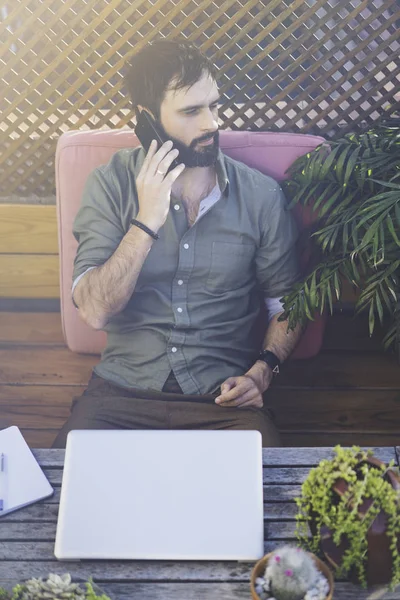 Uomo hipster che lavora sul computer portatile e utilizzando il telefono cellulare mentre seduto sulla terrazza del caffè. Attraente uomo barbuto che chiama ad amici.Bokeh . — Foto Stock