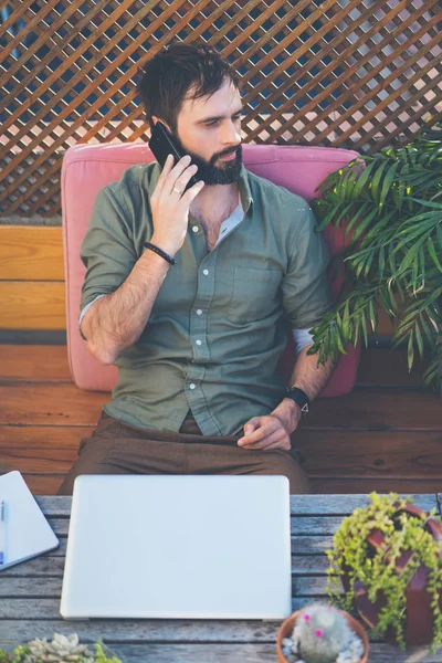 Uomo hipster che lavora sul computer portatile e utilizzando il telefono cellulare mentre seduto sulla terrazza del caffè. Attraente uomo barbuto che chiama ad amici.Verticale . — Foto Stock