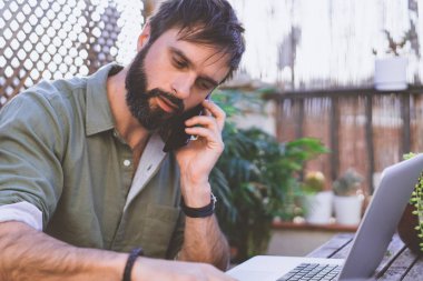 Dizüstü bilgisayarda çalışarak ve kafe Teras cep telefonunuzun vintage doğal kaba ahşap masasına oturan sakallı adam etrafında yeşil flores ve kaktüs. İşyeri dışında kavram çalışması.
