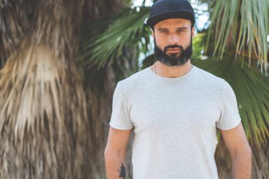 Hipster gri boş tişört ve siyah snapback kapağı için logo veya tasarım alanı ile rahat kentsel tarzı giymiş sakallı yakışıklı erkek modeli. Yeşil palmiye ve kaktüs özgeçmişlerine Bahçe.