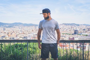 Hipster gri boş tişört ve siyah snapback kapağı için logo veya tasarım alanı ile rahat kentsel tarzı giymiş sakallı yakışıklı erkek modeli. Modern Bcn şehir panoramik görünümü arka planda.