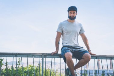 Sakallı kas hipster man modeli gri boş tişört ve siyah beysbol şapkası için logo veya tasarım alanı ile rahat spor kentsel tarzı giyiyor. Modern şehir panoramik görünümü arka planda.