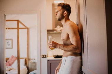 Seksi genç adam el portakal suyu tutarak, pencereden dışarı bakarak ve evde mutfakta dururken gülümseyen çıplak gövde ile