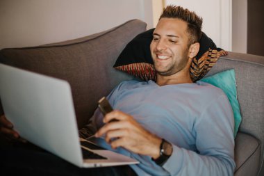 Evde çalışma anlar. Rahat kıyafetler modern daire kanepe laptop üzerinde genç beyaz birlikte çalışan adam. Arka plan bulanık.