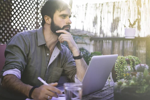 Attraktiv Bearded Man sitter vid vintage naturliga grov trä skrivbord arbetar på bärbar dator på café terrass omgivna gröna flores och cactus. Kontoret arbeta koncept. — Stockfoto