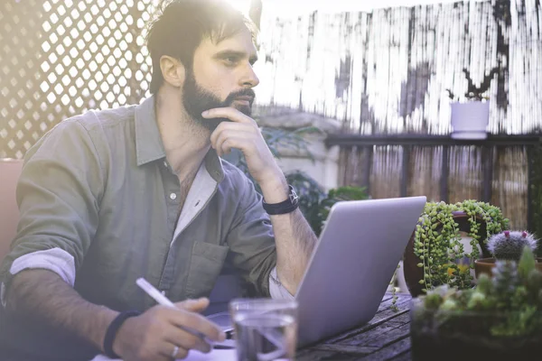 Självsäker Bearded Man sitter vid vintage naturliga grov trä skrivbord arbetar på bärbar dator på café terrass omgiven grön flores och cactus. Kontoret arbeta koncept. — Stockfoto