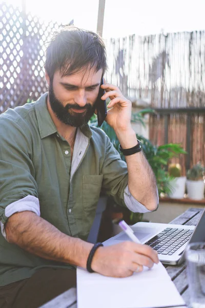 Uomo barbuto seduto alla scrivania vintage in legno grezzo naturale che lavora sul computer portatile e utilizza il telefono cellulare sulla terrazza del caffè circondato da flora e cactus verdi.Fuori dal concetto di lavoro d'ufficio. — Foto Stock