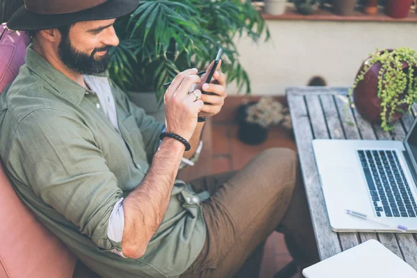 Skäggiga säker hipster bär grön skjorta och bruna hatt SMS meddelande via smartphone på terrassen utanför. Positiv man avkopplande på sommardag. — Stockfoto