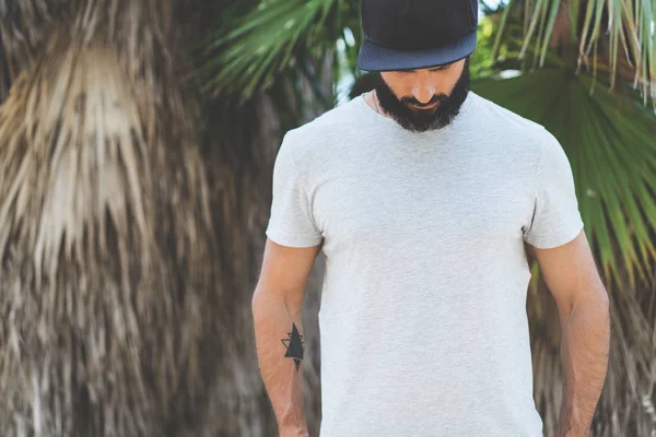 Bearded muscular hipster man model wearing gray blank t-shirt and a black baseball cap with space for your logo or design in casual urban style.Green palm and cactus garden on the background.