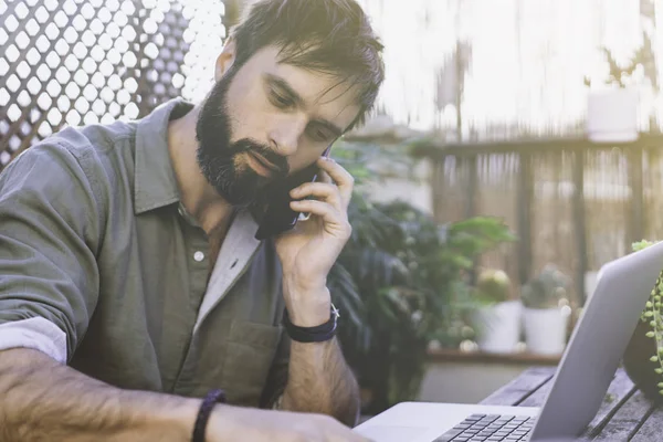 Uomo barbuto seduto alla scrivania in legno ruvido naturale vintage che lavora sul computer portatile e utilizza il telefono cellulare sulla terrazza del caffè circondato da flore verdi e cactus. . — Foto Stock