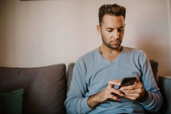 Young handsome man sitting at home on sofa and using mobile phone. Men holding smartphone hands and typing text message.