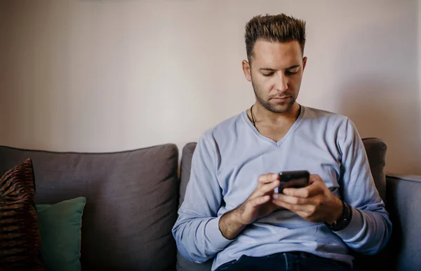 Stilig ung man sitter hemma på soffan och med hjälp av mobiltelefon. Män hålla smartphone händer och skriva SMS. — Stockfoto