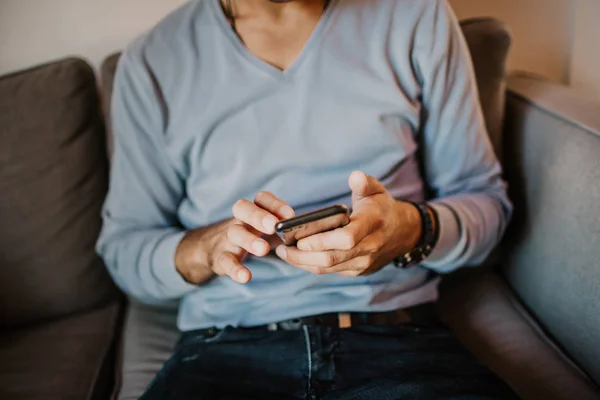 Junger gutaussehender Mann, der zu Hause auf dem Sofa sitzt und sein Handy benutzt. Männer halten Smartphone in der Hand und tippen SMS. — Stockfoto