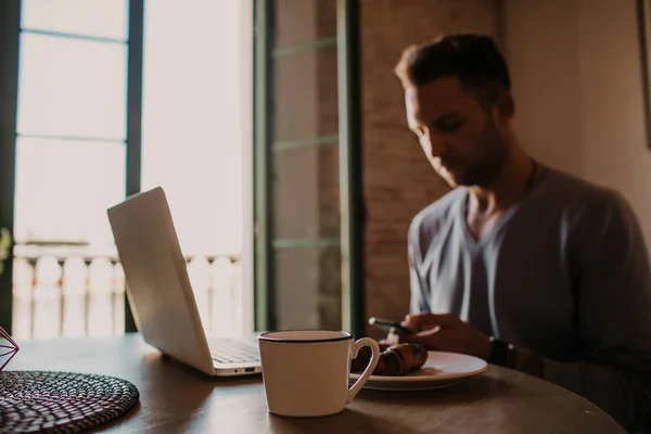 Stilig medarbetare man som arbetar på vardagsrum hemma. Man sitter på träbord med laptop och mobiltelefon. Suddig bakgrund. — Stockfoto