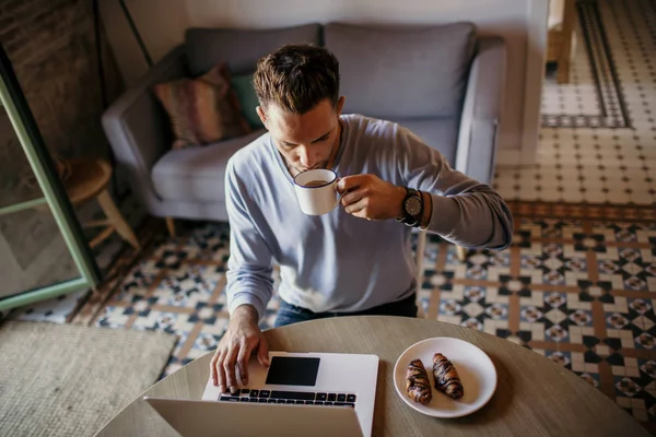 Bonito colega de trabalho a trabalhar na sala de estar em casa. Homem sentado à mesa de madeira usando laptop e telefone celular. Fundo desfocado . — Fotografia de Stock