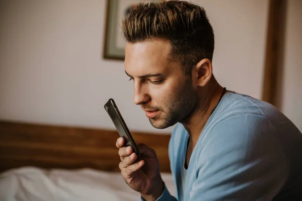 Bonito homem usando smartphone móvel para navegar na internet e enviar mensagem de áudio. Homem usando gadget no apartamento moderno . — Fotografia de Stock