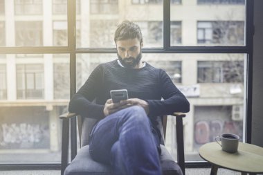 Cep telefonu uygulaması Mobil blogging için kullanarak siyah kazak beyaz adam sakallı. Kendine güvenen genç hipster adam modern loft, sosyal ağ Haberler okuyabilir. Arka plan bulanık. Mercek parlamaları.