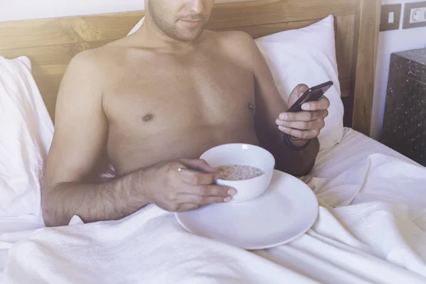 Handsome muscular male gay lying on bed with smartphone, healthy breakfast in the morning at bedroom.Shirtless sexy man enjoying new day