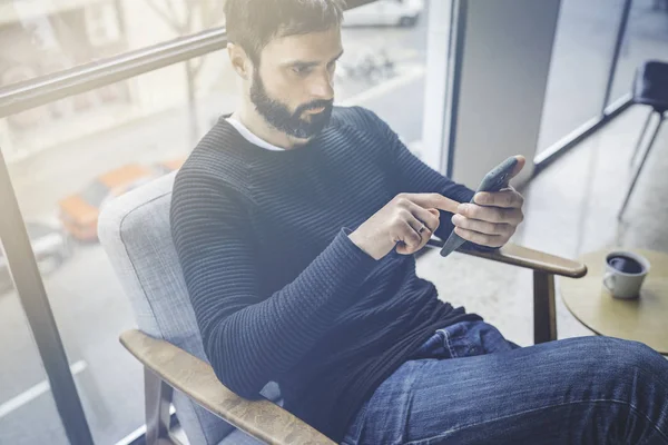 Guapo barbudo hombre caucásico en jersey negro usando aplicación de teléfono móvil para blogging móvil. Confiado joven hipster chico leer noticias de las redes sociales en loft.Blurred moderna background.Flares . —  Fotos de Stock