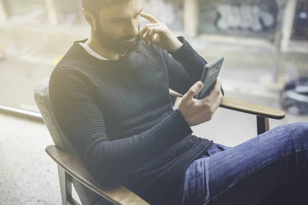 Homem hispânico barbudo alegre em pulôver preto usando o aplicativo de telefone celular para blogs móveis. Confiante jovem hipster cara ler notícias da rede social no moderno loft.Blurred background.Flares . — Fotografia de Stock