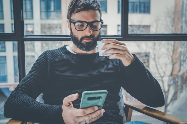 Skäggig spansktalande mannen i svart tröja med mobiltelefon ansökan för mobil blogg. Självsäker ung hipster guy Läs nyheter via mobiltelefon från sociala nätverk på moderna loft. Suddig bakgrund. — Stockfoto
