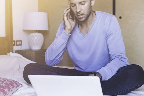 Stilig medarbetare man som arbetar på vardagsrum hemma. Man sitter på sängen med laptop och mobiltelefon. Suddig bakgrund. Nödraketer. — Stockfoto