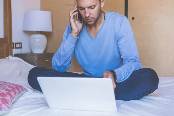 Jeune collègue attrayant travaillant dans le salon à la maison. Homme assis au lit en utilisant un ordinateur portable et un téléphone mobile. Fond flou — Photo