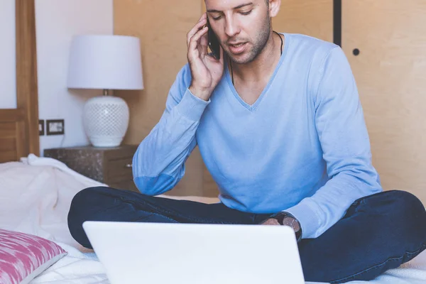 Självsäkra unga medarbetare man som arbetar på vardagsrum hemma. Man sitter på sängen med laptop och mobiltelefon. Suddig bakgrund — Stockfoto