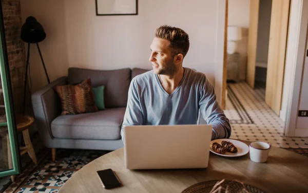 Stilig medarbetare man som arbetar på vardagsrum hemma. Man sitter på träbord med laptop under frukosten på kök. Suddig bakgrund. — Stockfoto