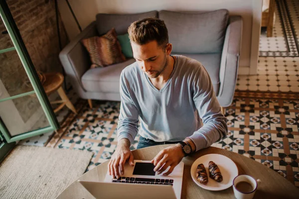 Stilig medarbetare man som arbetar på vardagsrum hemma. Man sitter på träbord med laptop under frukosten på kök. Suddig bakgrund. — Stockfoto
