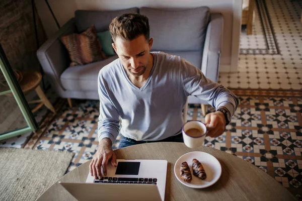 Stilig medarbetare man som arbetar på vardagsrum hemma. Man sitter på träbord med laptop under frukosten på kök. Suddig bakgrund. — Stockfoto