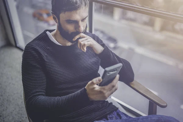 Bearded hispanic man in black pullover using mobile phone application for mobile blog. Confident young hipster guy read news via cellphone from social network at modern loft.Blurred background.Flares. Stock Picture