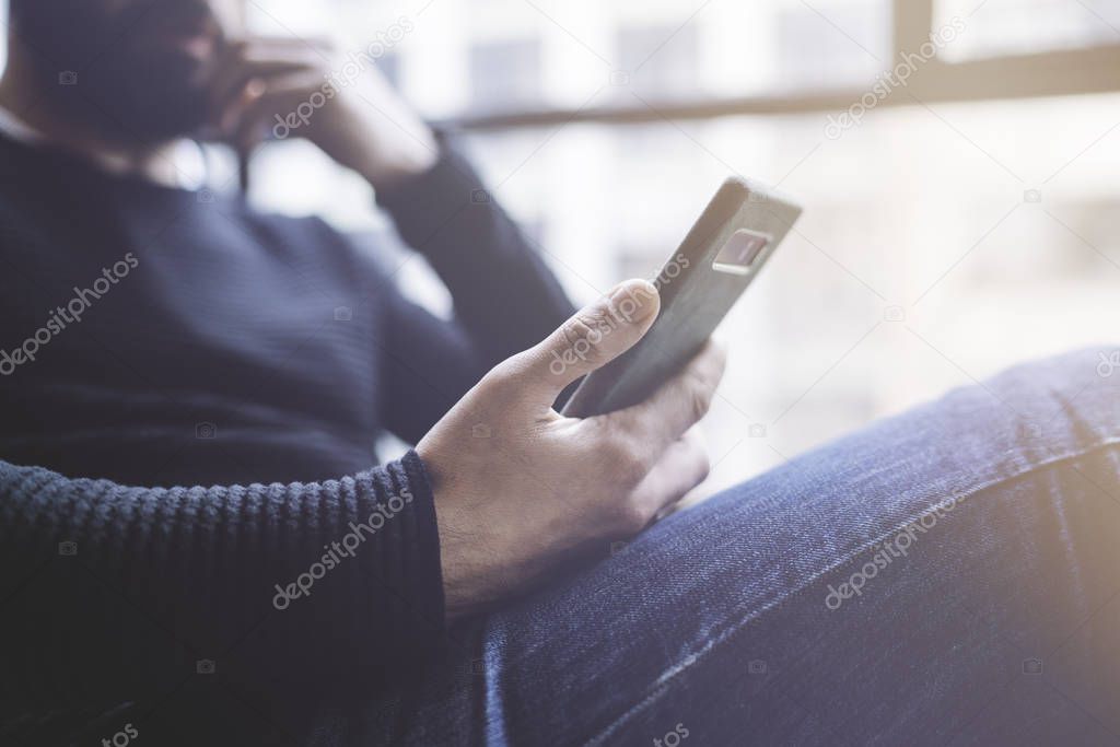 Bearded hispanic man in black pullover using mobile phone application for mobile blog. Confident young hipster guy read news via cellphone from social network at modern loft.Blurred background.