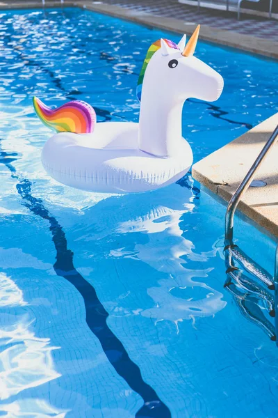 Надувні барвисті білий єдиноріг біля басейну. Відпустка у басейні плавають з Пластмасові іграшки. Концепція релаксації і веселощів. Брижі води в басейні зі відображення сонця. Вертикаль. — стокове фото
