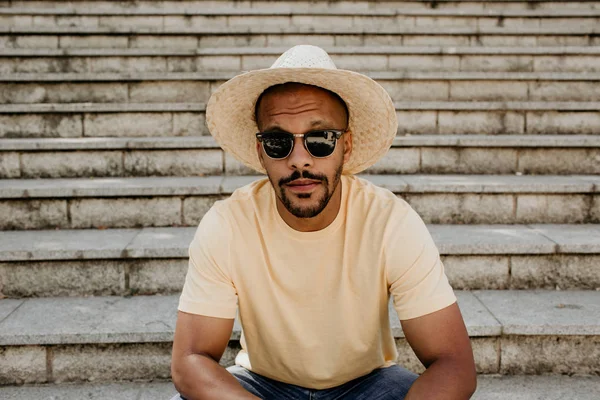 Stijlvolle donkere man in trendy strooien hoed en zonnebril zittend op de trap. Afrikaanse hipster treveler ontspannen in city. Ontspanning en rust bij zomerdag. — Stockfoto