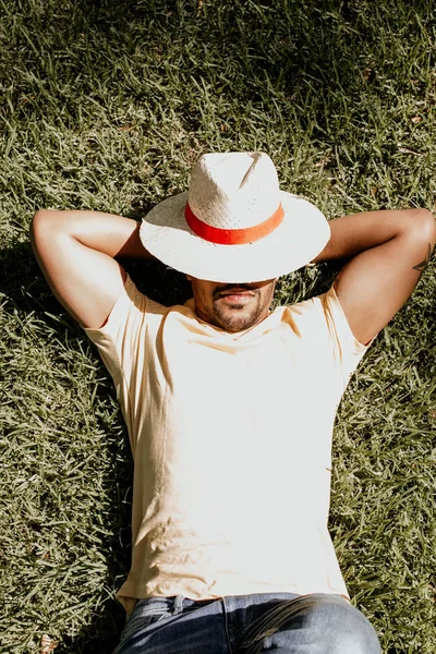 African-American male model laying in the green grass with trendy straw hat.African man hipster treveler relaxing at park. Leisure and rest at summer day.Vertical.