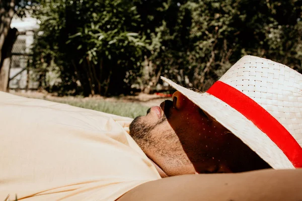 Portrait of African-American male model laying in the green grass with trendy straw hat.African man hipster treveler relaxing at park. Leisure and rest at summer day.