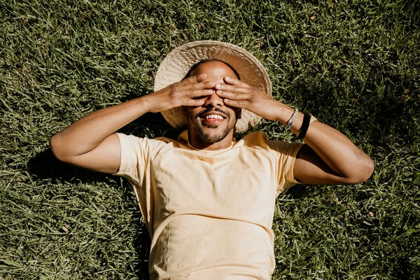 African-American male model laying in the green grass with trendy straw hat and closed eyes by hands.African man hipster treveler relaxing at park. Leisure and rest at summer day.