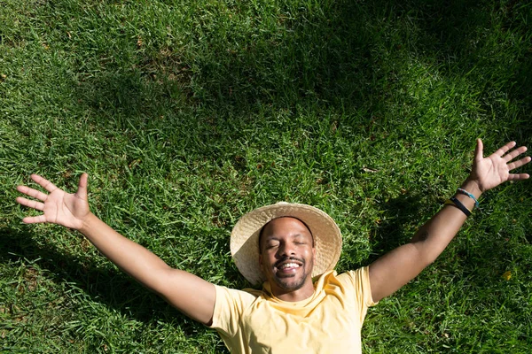 Happy African-American male model laying in the green grass with trendy straw hat and spread his hands.African man hipster treveler relaxing at park. Leisure and rest at summer day.