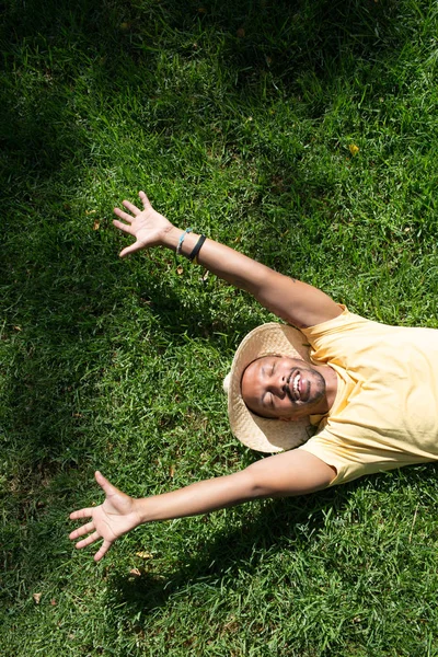 Happy African-American male model laying in the green grass with trendy straw hat and spread his hands.African man hipster treveler relaxing at park. Leisure and rest at summer day.Vertical.