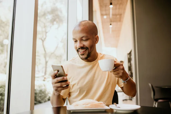 Fröhlich fröhlicher afrikanischer Hipster hält Tasse in der Hand, trinkt frischen Cappuccino, surft im Internet und checkt den Newsfeed in den sozialen Medien.. — Stockfoto