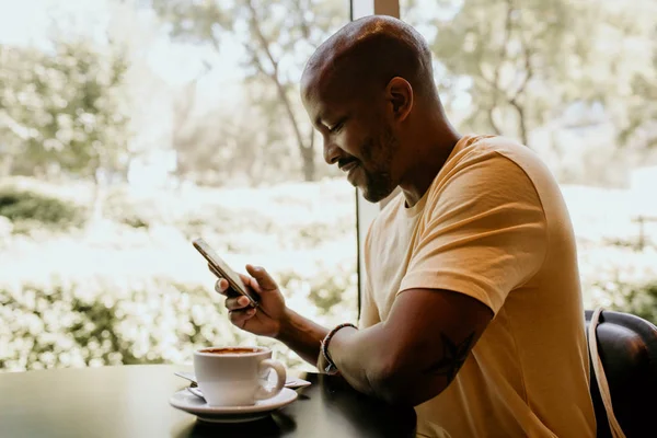 Feliz hipster africano alegre celebración taza, beber capuchino fresco, navegar por Internet y comprobar noticias en las redes sociales. Hombre usando el teléfono celular durante el descanso de café en la cafetería moderna . —  Fotos de Stock