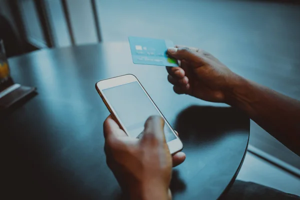 Vértes kilátás mozgatható telefon megható férfi kezét. Afrikai férfi smartphone és hitelkártya segítségével a fából készült asztalnál ülve. Üres fehér üres másol hely képernyő adatait. Makett. — Stock Fotó