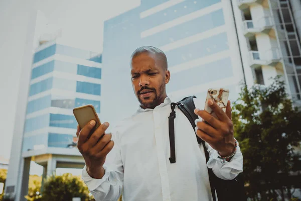 Positiv afrikanisch-amerikanischer Rüde mit zwei Mobiltelefonen im Freien. Bürohochhäuser im Hintergrund. Geschäftsstress und hartes Arbeitskonzept. — Stockfoto