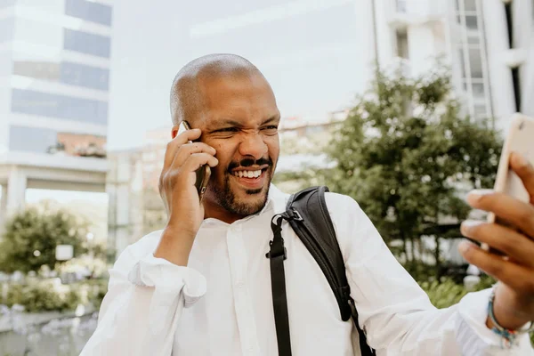 Homme afro-américain positif utilisant deux téléphones portables en plein air. Des gratte-ciel de bureau en arrière-plan. Stress d'affaires et concept de travail acharné . — Photo