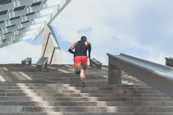 Musculoso hispano Joven atleta de piel oscura corriendo por un tramo de escaleras con velocidad, joven deportivo en entrenamiento de camiseta negra o haciendo ejercicio al aire libre mientras trota por los escalones. Vista trasera . —  Fotos de Stock