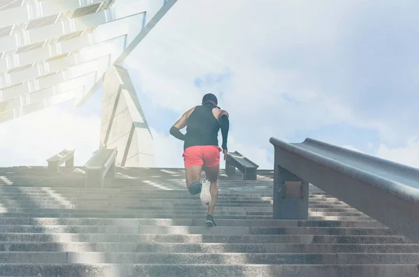 Musculoso caucásico Joven atleta de piel oscura corriendo por un tramo de escaleras con velocidad, joven deportivo en entrenamiento camiseta negra o haciendo ejercicio al aire libre mientras trota por los escalones. Vista trasera . —  Fotos de Stock