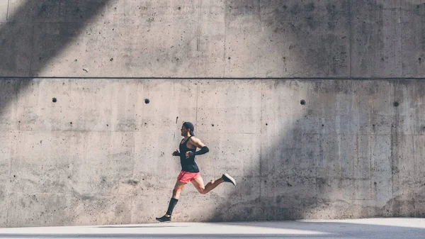 Hombre musculoso atleta velocista corriendo rápido, haciendo ejercicio al aire libre, trotando fuera contra fondo de hormigón gris con área de espacio de copia para el mensaje de texto o el contenido del anuncio.Vista lateral, longitud completa . —  Fotos de Stock